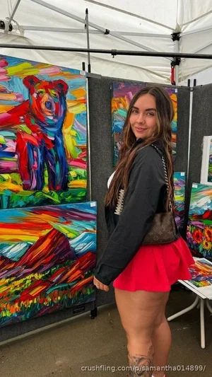 Woman in black top and red skirt poses at art market with colorful paintings behind her.