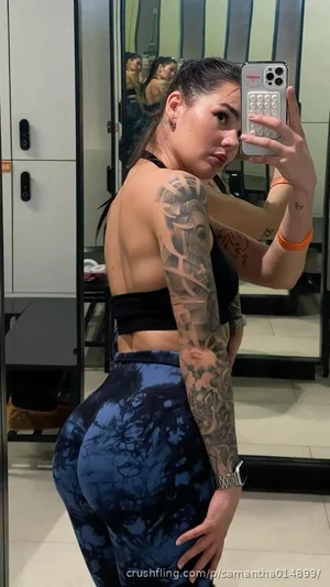Woman with tattoos in black sports bra and blue leggings takes mirror selfie in indoor setting.