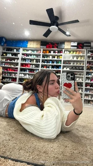 Woman in white sweater and blue top lying on carpet, taking mirror selfie in room with shoe shelves.