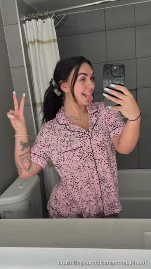 Woman in pink patterned pajama shirt taking mirror selfie in bathroom, tongue out, peace sign.