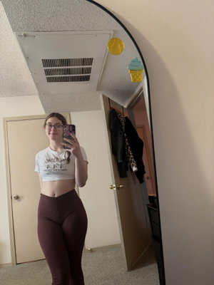 Woman in white crop top and brown leggings takes mirror selfie in bedroom.