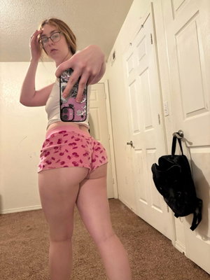 Woman in white crop top and pink shorts taking mirror selfie in hallway.