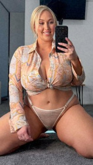 Blonde woman in patterned shirt and sheer underwear kneeling indoors for mirror selfie.