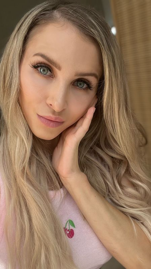 Woman with long blonde hair and blue eyes wearing pink top with cherry design, posing indoors.