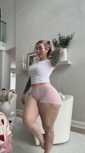 Woman with blonde hair and tattoos wears white crop top and pink shorts in a modern living room.