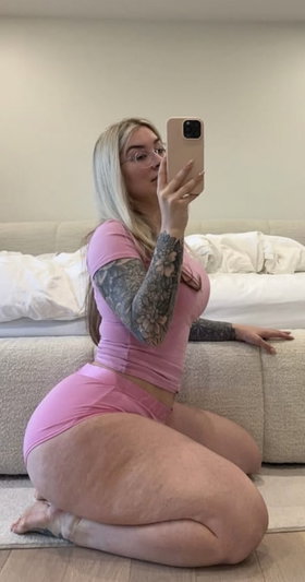 Woman with blonde hair and tattoos wears pink outfit, kneeling on floor in front of couch.