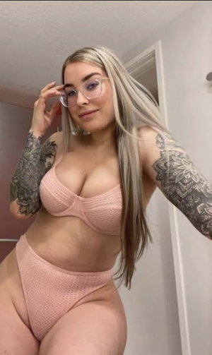 Woman with long blonde hair and tattoos wears pink lingerie, posing indoors.