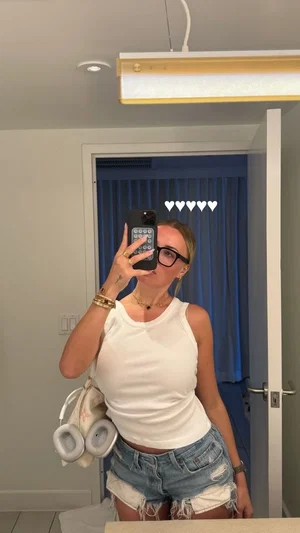 Woman in white tank top and denim shorts taking mirror selfie in bedroom.