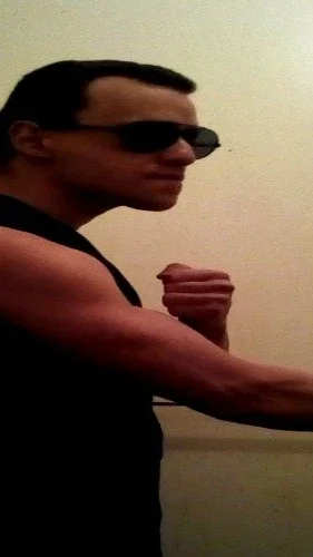 Man in black tank top flexing arm muscles indoors.