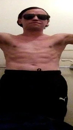 Shirtless man wearing sunglasses and black sweatpants, standing indoors.