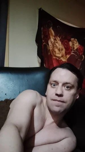 Shirtless man with dark hair lying on a dark surface, floral curtain in background.