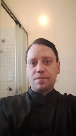 Man in black shirt taking selfie in bathroom with tiled shower visible.