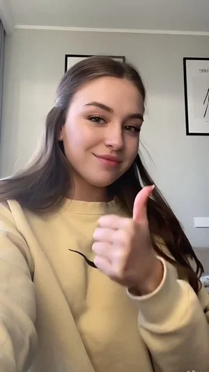 Woman with long brown hair in a beige sweatshirt giving a thumbs-up indoors.