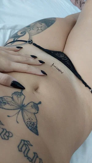 Woman with tattoos on stomach and side, wearing black lace underwear, lying on white sheets.