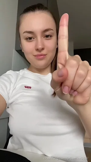 Woman in white t-shirt pointing finger at camera indoors.