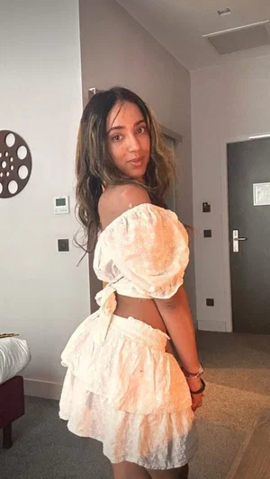 Woman in off-shoulder white dress posing indoors, long wavy hair, bedroom setting.