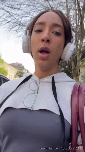 Woman in gray and white hoodie with headphones, outdoors with trees and buildings.