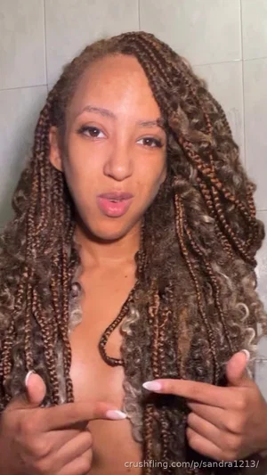 Woman with long braided hair pointing at her chest, bare skin visible, indoor setting.