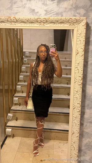 Woman with long braids in white crop top and black skirt, taking mirror selfie in marble staircase.