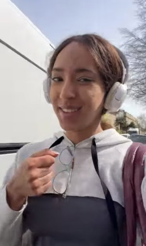 Woman with headphones and glasses, wearing a gray and white hoodie, outdoors near a white van.
