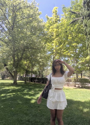 Woman in white crop top and skirt standing in a grassy park with trees.