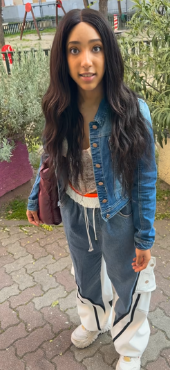 Woman with long dark hair in denim jacket and sweatpants, standing outdoors near plants.