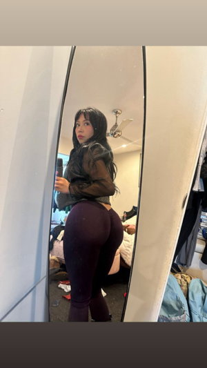 Woman in sheer top and dark leggings poses in mirror selfie in bedroom.