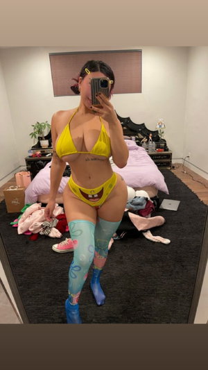 Woman in yellow bikini and blue thigh-high socks poses in bedroom mirror selfie.