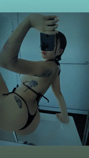 Woman in black lingerie with tattoos takes mirror selfie in bathroom.