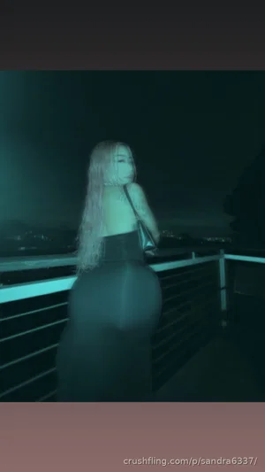 Woman with long blonde hair in dark outfit standing on balcony at night.