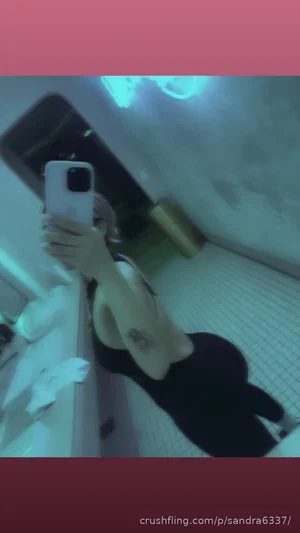 Woman in black top and pants taking mirror selfie in bathroom with tiled floor.