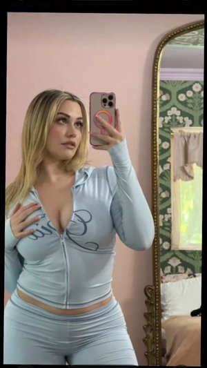 Blonde woman in blue zip hoodie and pants in bedroom mirror selfie.