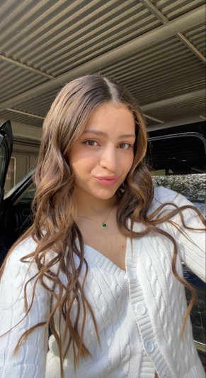 Woman with long wavy hair in white cardigan, standing near car under metal roof.