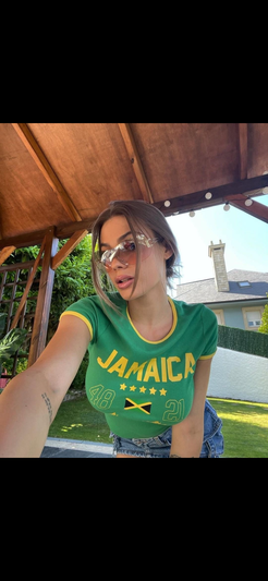 Woman in green Jamaica t-shirt and denim shorts, taking selfie under wooden pergola outdoors.