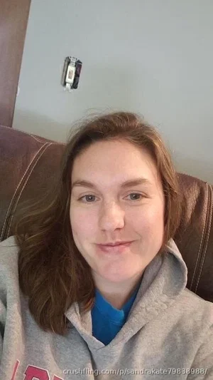 Woman with brown hair wearing gray hoodie and blue shirt, sitting on brown couch indoors.
