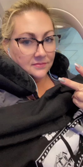Blonde woman with glasses wearing black jacket taking selfie indoors.