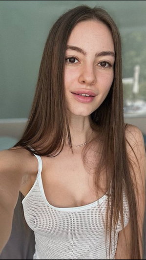 Woman with long brown hair in white mesh top taking selfie indoors.
