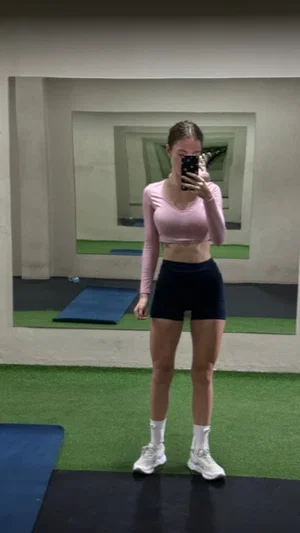 Woman in pink crop top and black shorts taking mirror selfie in gym.