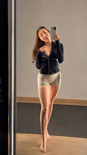 Woman in black zip-up top and beige shorts takes mirror selfie indoors.