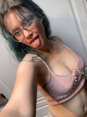 Blue-haired woman with glasses in pink top taking selfie indoors.