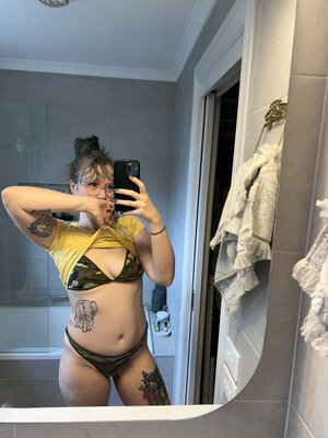 Woman in camo bikini and yellow top, tattoos, mirror selfie in bathroom.