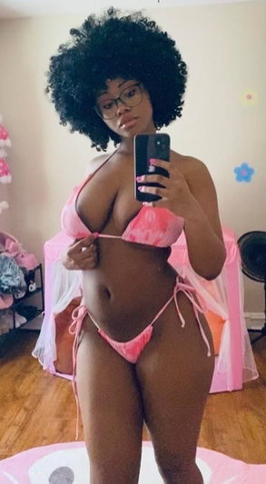Curvy woman with afro hair and glasses in pink bikini taking mirror selfie.