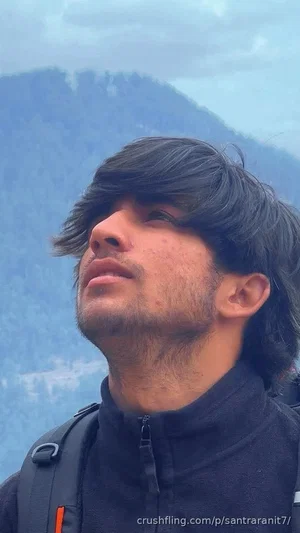 Man with dark hair and beard wearing a black jacket, standing outdoors with mountains in the
