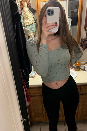 Woman in floral crop top and black leggings taking mirror selfie in bathroom.