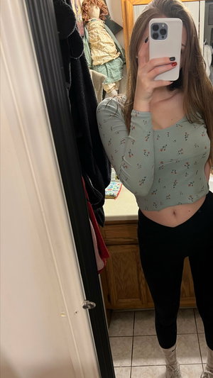 Woman in floral crop top and black leggings taking mirror selfie in room.