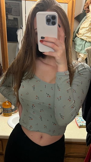 Woman in floral crop top and black pants taking mirror selfie in bathroom.