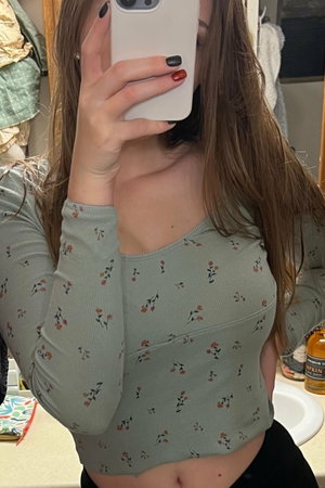 Woman with long brown hair in floral crop top taking mirror selfie in bathroom.
