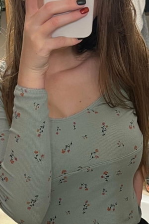 Woman with long brown hair wearing floral long-sleeve top, holding phone to face in indoor setting.