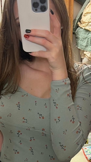Woman with long brown hair in floral long-sleeve top taking mirror selfie indoors.