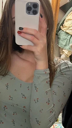 Woman with long brown hair in floral top taking mirror selfie indoors.